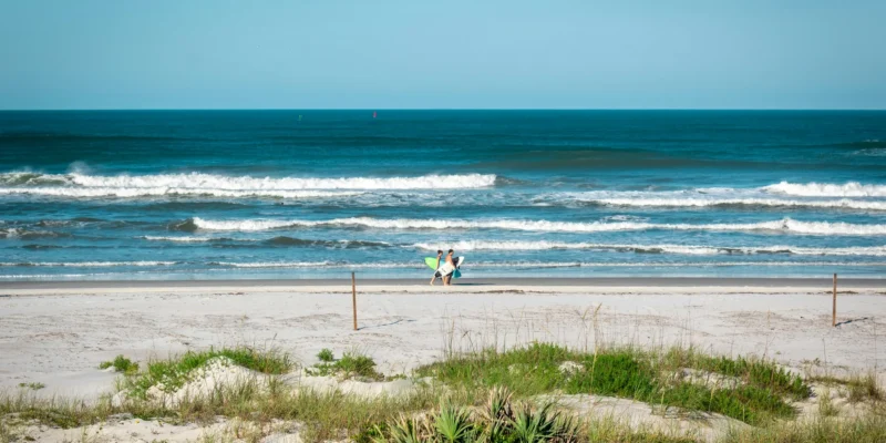 Zwei Menschen mit Surfbrettern am Strand von New Smyrna Beach, dahinter die Brandung