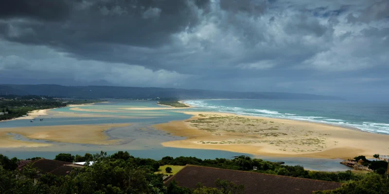Blick über Lagune, Strand und Ozean bei Plettenberg Bay unter dunklen Sturmwolken